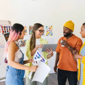 future search: A diverse group of coworkers engage in a creative discussion around a business plan. They review color samples and notes, with a whiteboard displaying the plan and action steps in the background.