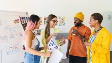 future search: A diverse group of coworkers engage in a creative discussion around a business plan. They review color samples and notes, with a whiteboard displaying the plan and action steps in the background.